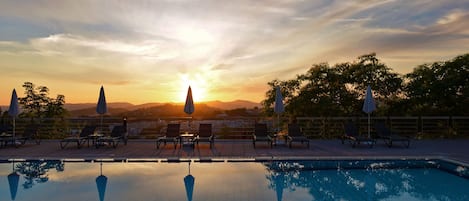 Zona junto a la piscina con sillones de descanso y sombrillas, reflejando el atardecer y montañas al fondo.