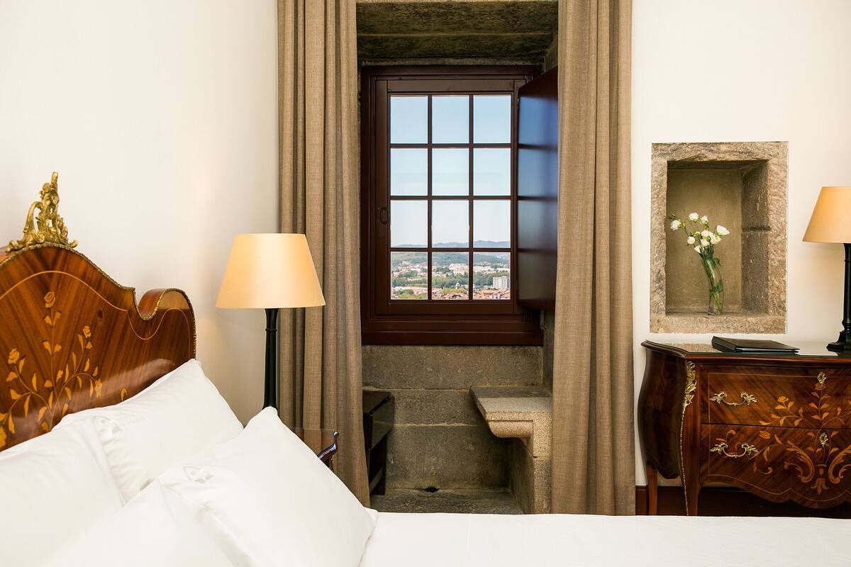 Une chambre d’hôtel avec un lit, un bureau, une lampe, une fenêtre donnant sur un paysage agréable et un petit escalier.