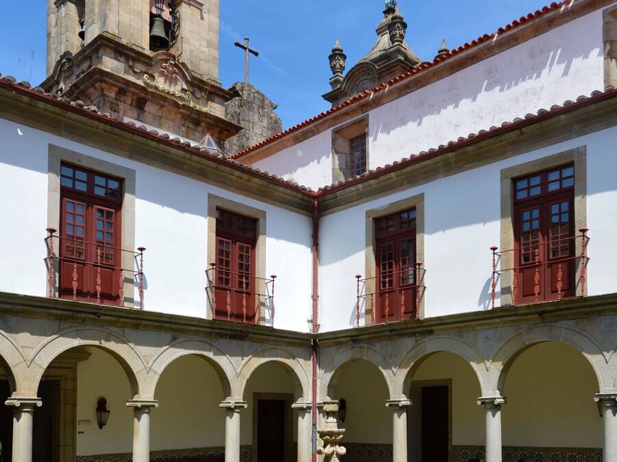 Un patio con fuente, paredes blancas, puertas rojas y una torre de campanas.