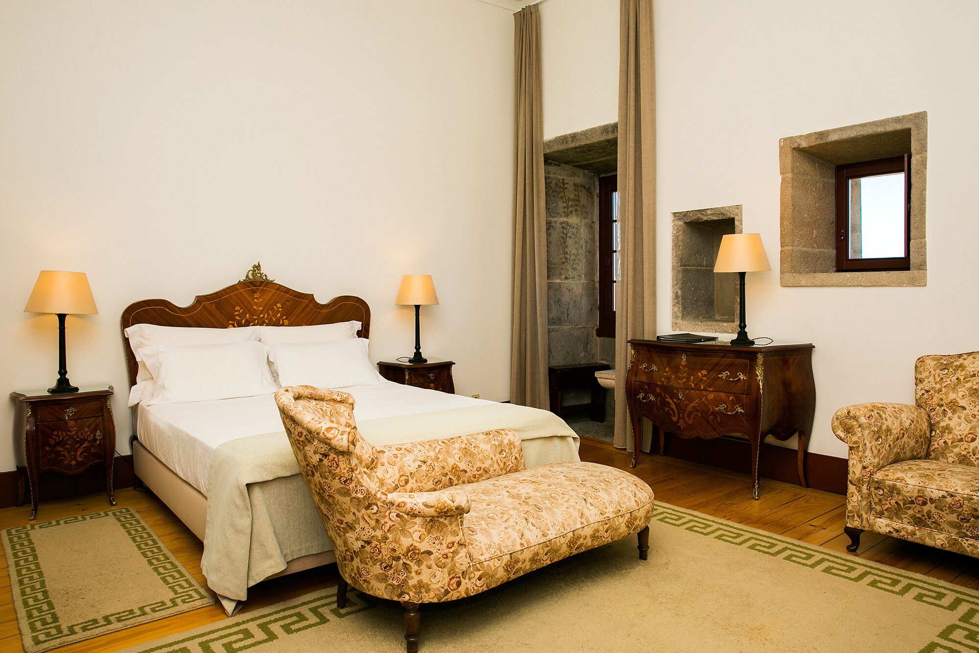 A hotel room with a bed, two chairs, a nightstand with lamps, a dresser, and a mirror.