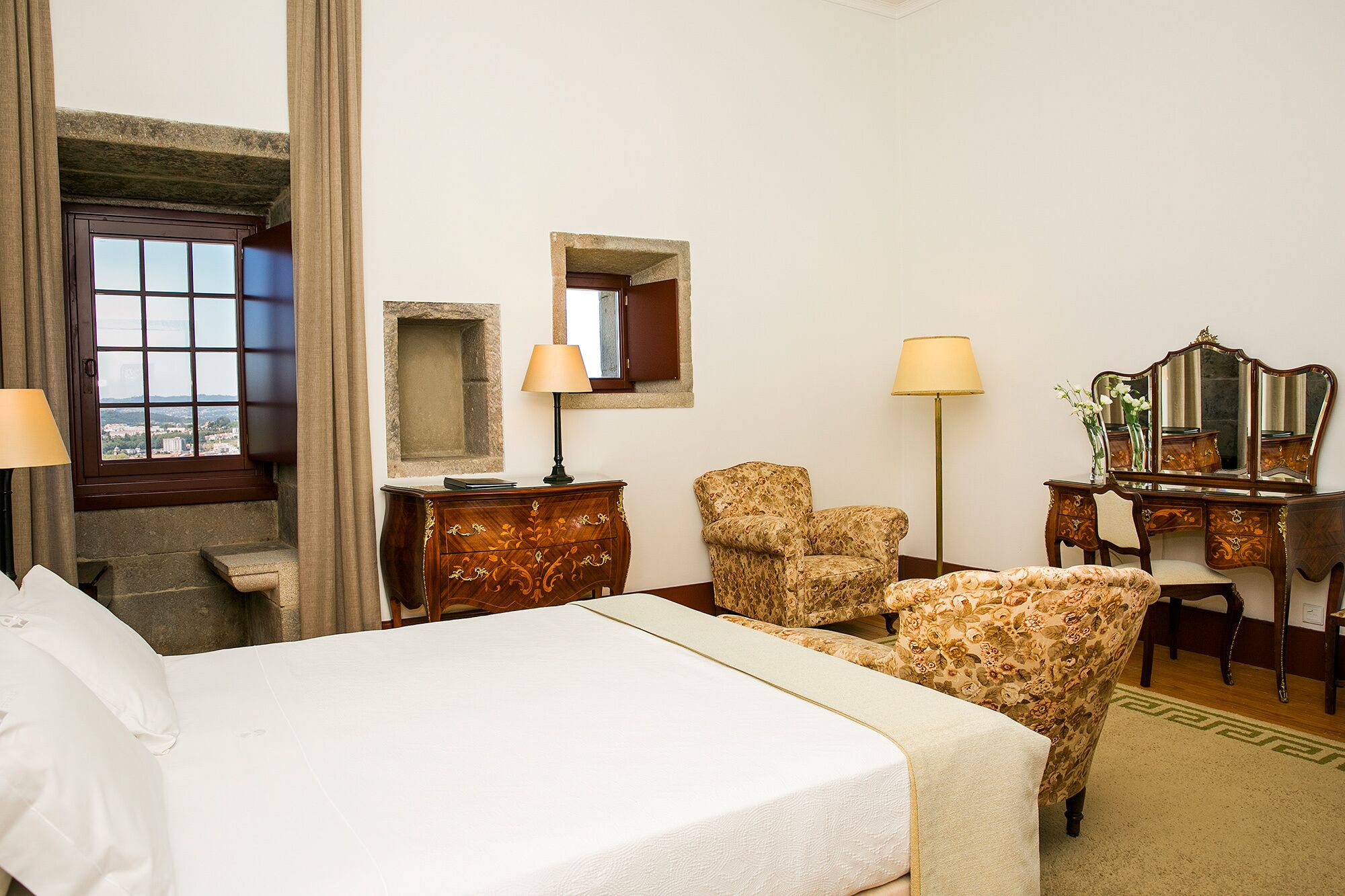 A hotel room with a bed, two chairs, a desk with a mirror, and a window with curtains.