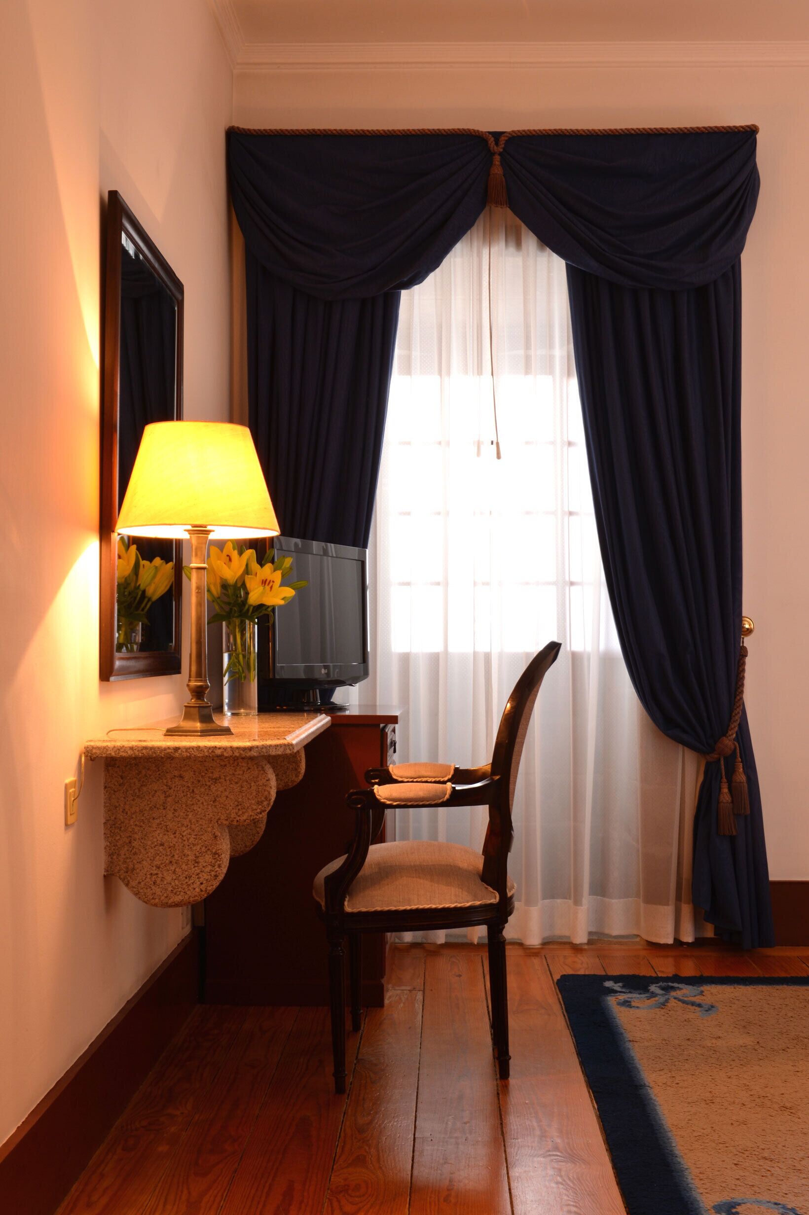 A hotel room with a wooden desk, a lamp, a chair, a television, and a window with dark curtains.