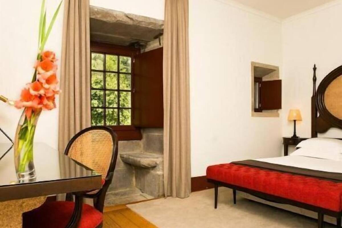 Une chambre d’hôtel avec un lit, une petite table et une chaise, un vase avec des fleurs et une fenêtre avec des rideaux.
