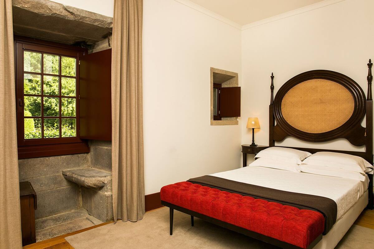 Une chambre avec un banc en pierre, un lit doté d’une tête de lit rembourrée en tissu rouge, un miroir rond et une fenêtre avec des rideaux.