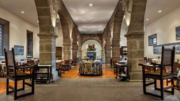 Un amplio salón con altas columnas de piedra, mesas y sillas de madera dispuestas para comer.
