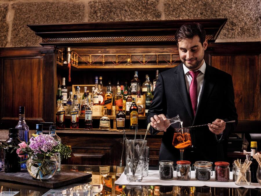 Un bar con un cantinero preparando una bebida, varias botellas y frascos con ingredientes, y una barra de madera.
