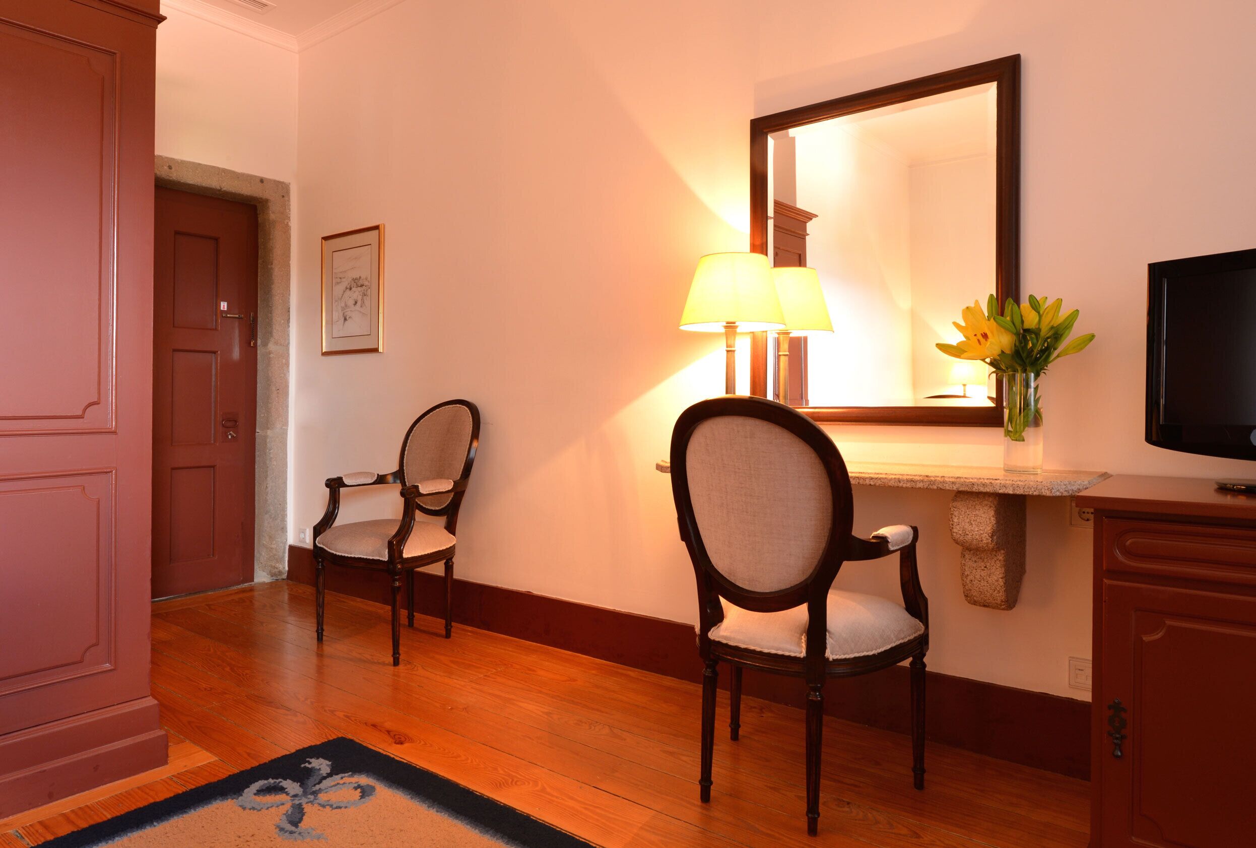 Habitación de hotel con piso de madera, dos sillones tapizados, un televisor, una lámpara, un espejo y un cuadro en la pared.