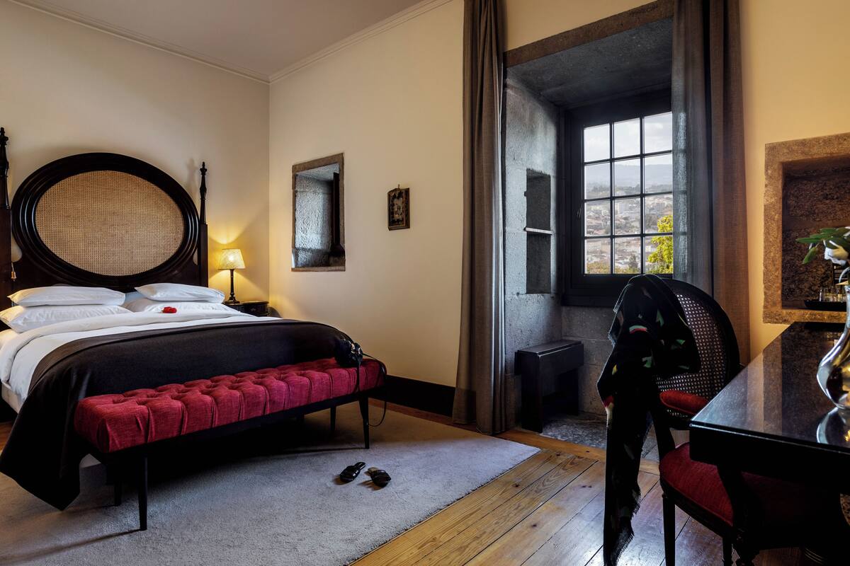 Une chambre avec un grand lit, un bureau avec une chaise, une fenêtre donnant sur un beau paysage et un vase rempli de fleurs posé sur le bureau.