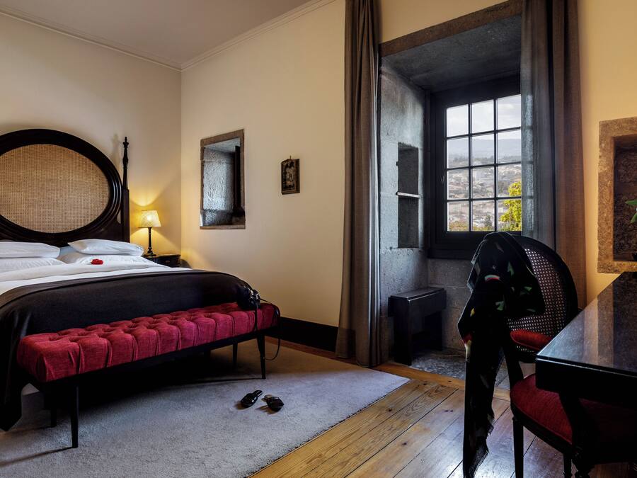 A bedroom with a large bed, a desk with a chair, a window with a view, and a vase with flowers on the desk.