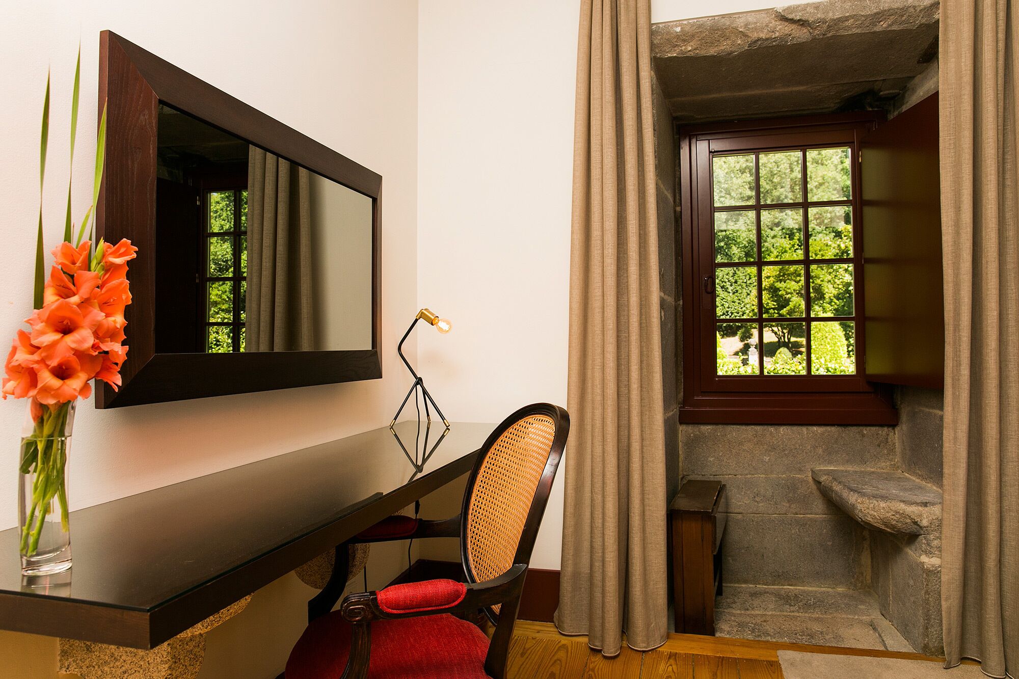 A bathroom with a wooden vanity, a mirror, a chair, a vase with flowers, and a window with curtains.