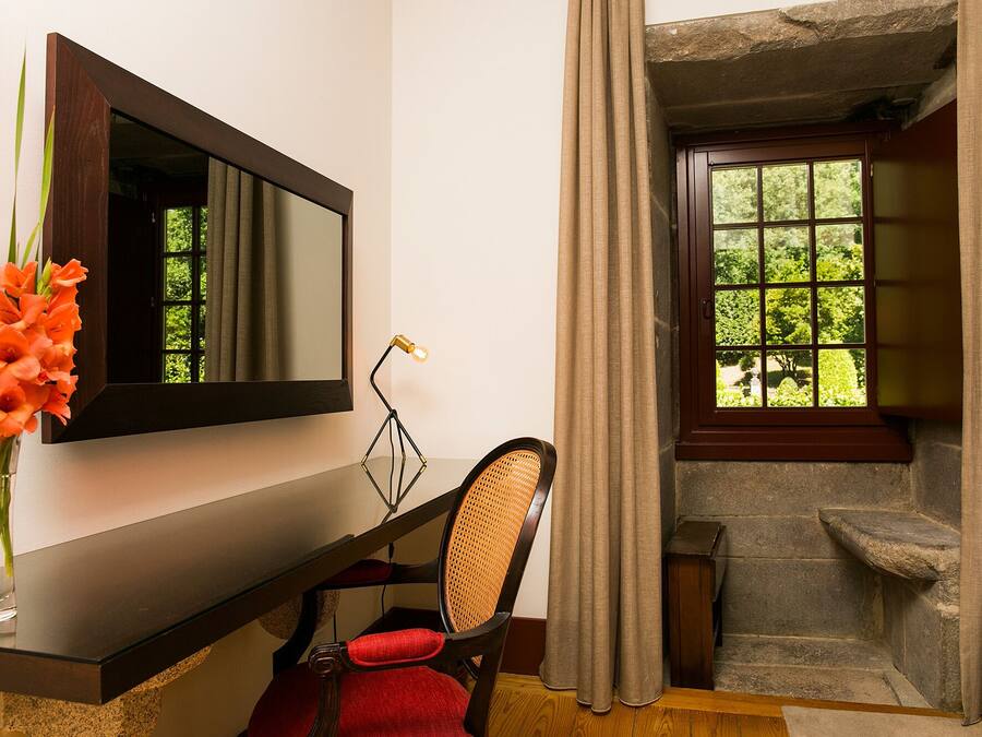 A bathroom with a wooden vanity, a mirror, a chair, a vase with flowers, and a window with curtains.