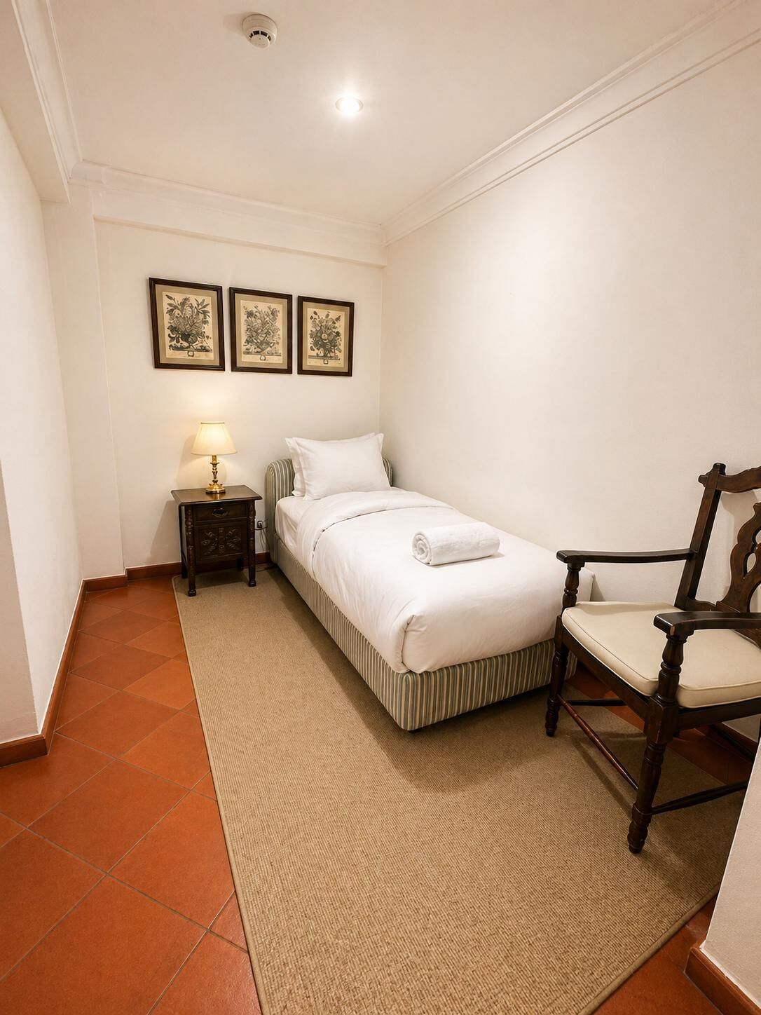 A single bed with a white comforter, a wooden nightstand with a lamp, a chair, and three framed botanical prints on the wall.