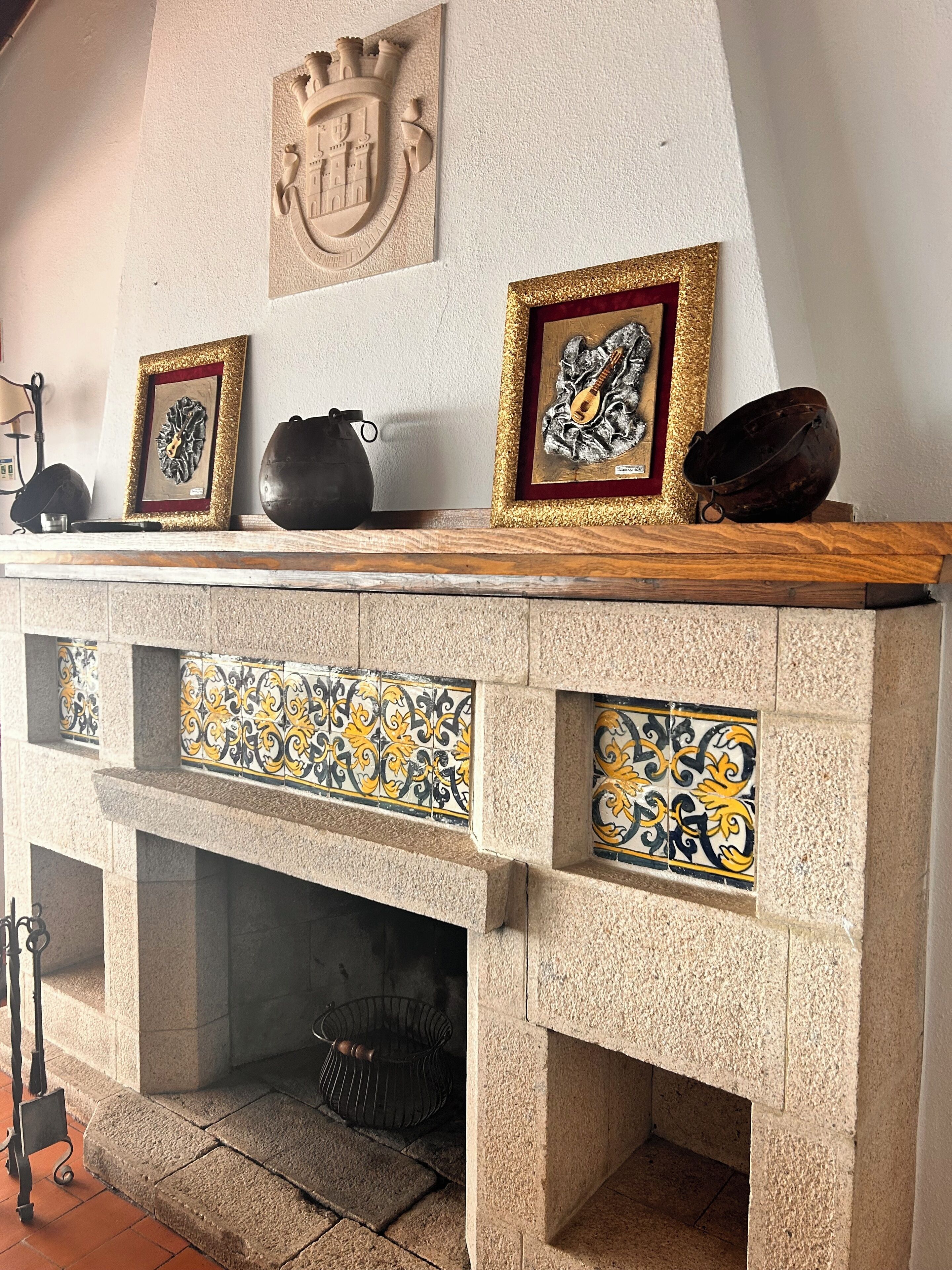 A fireplace with decorative tiles, a coat of arms, and framed artwork on the mantel.