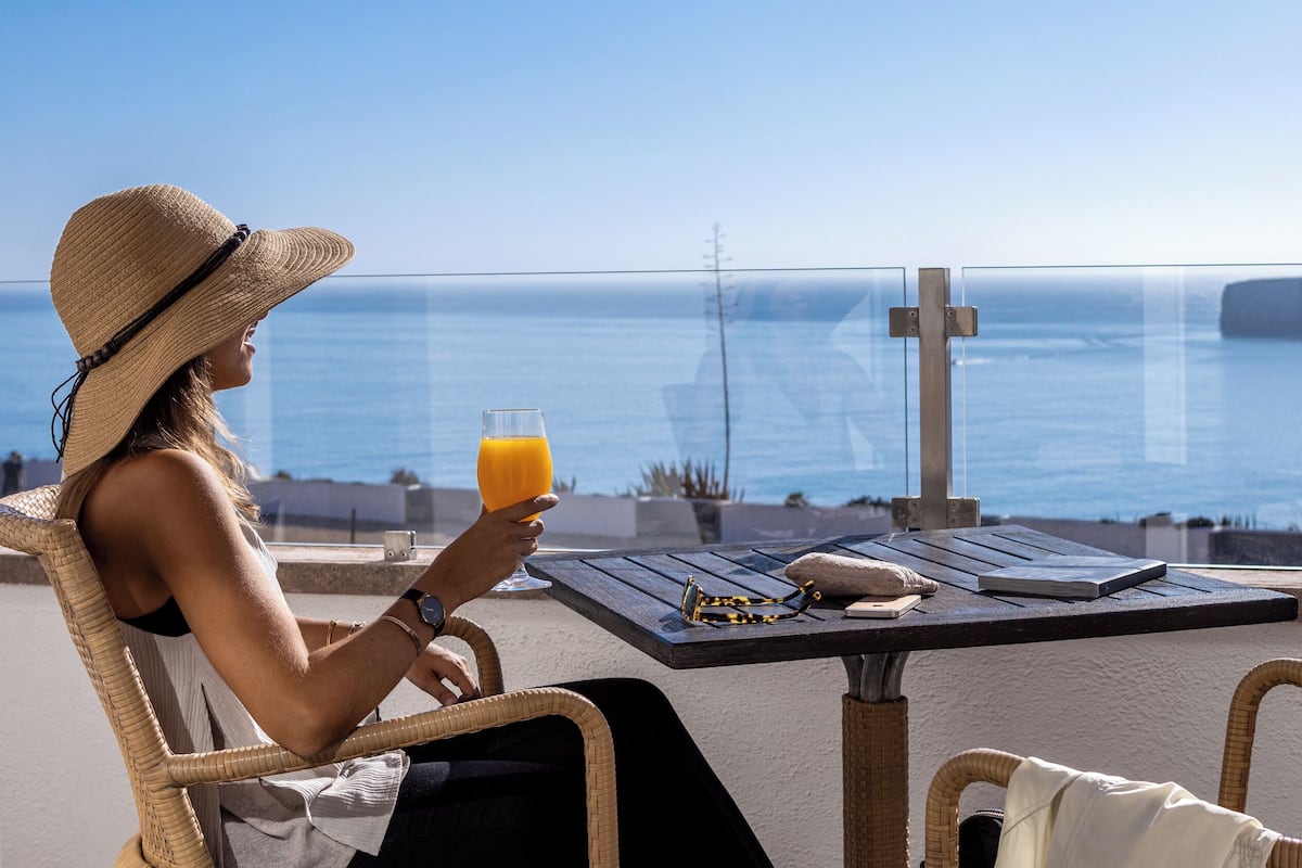 Eine Person sitzt an einem Tisch mit einem Orangensaftglas und blickt auf die Küstenlandschaft.