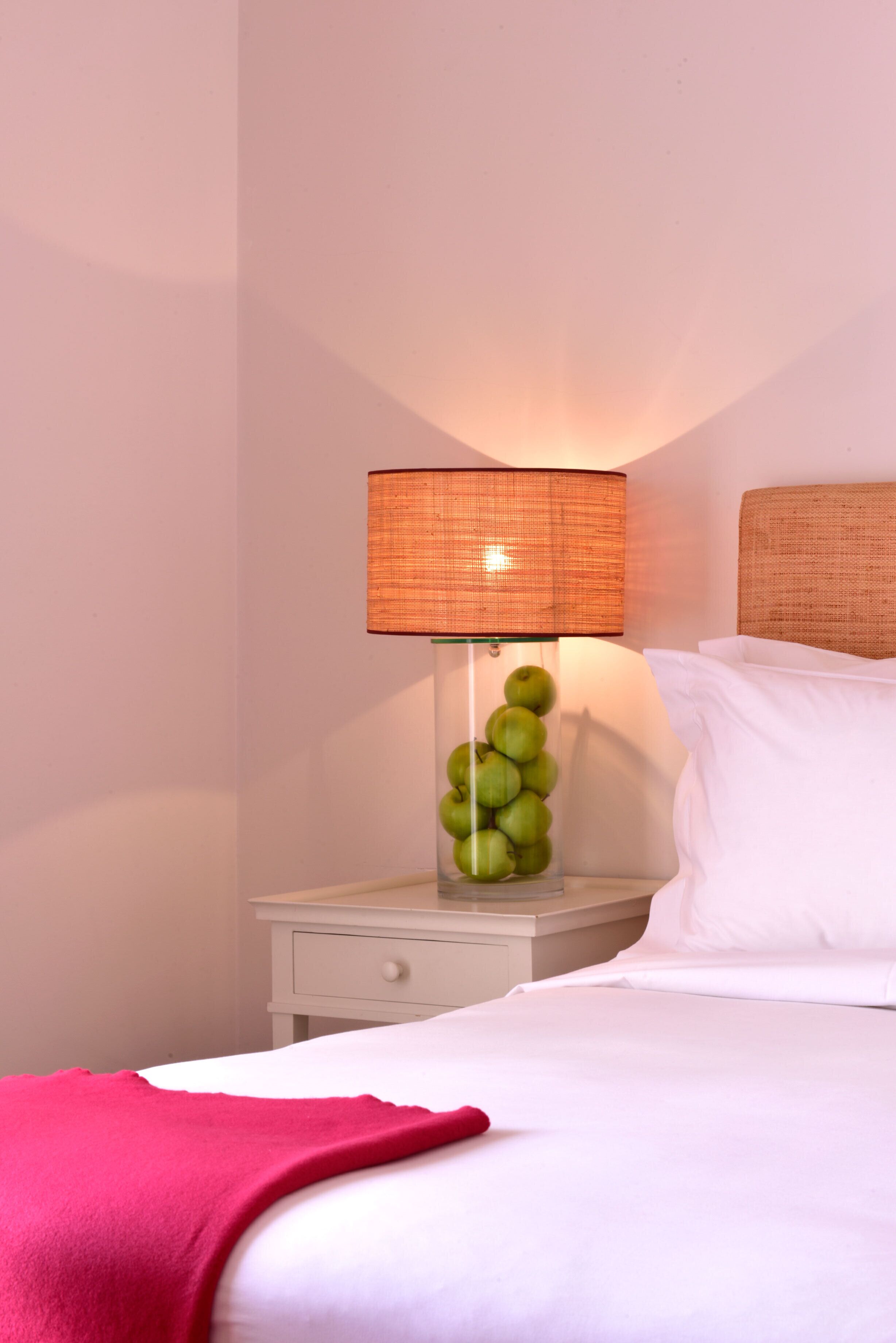 Een net opgemaakt bed met witte lakens, een rode plaid en een bedlamp met een gevlochten lampenkap.