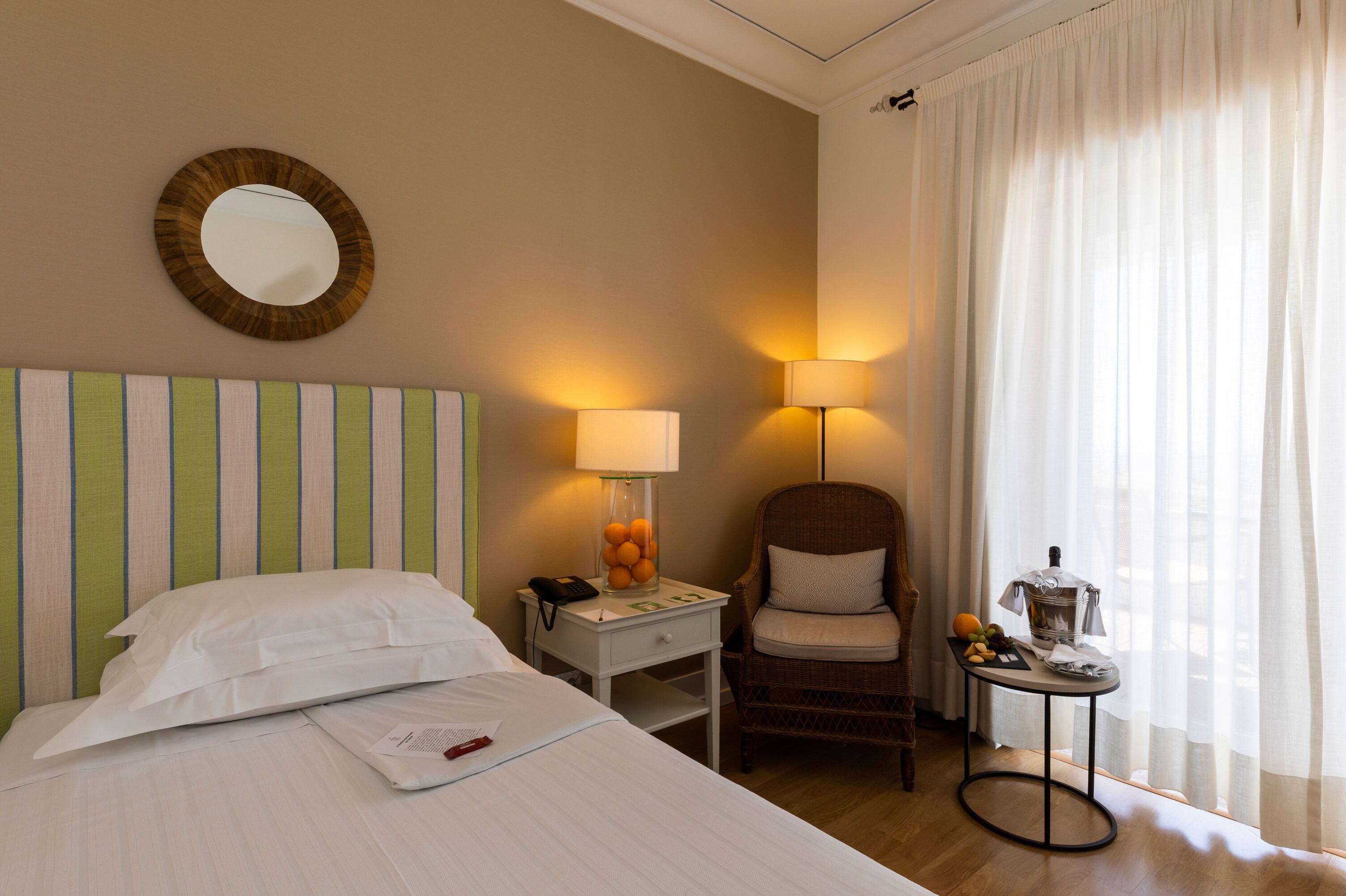 A hotel room with a bed, a chair, a small table with a lamp, and a window with curtains.