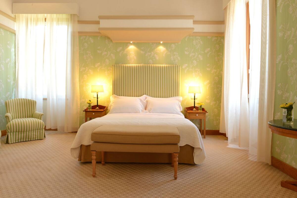 Habitació d'hotel amb un llit gran, dues taules de nit amb làmpades, una cadira i paper pintat amb motius florals.