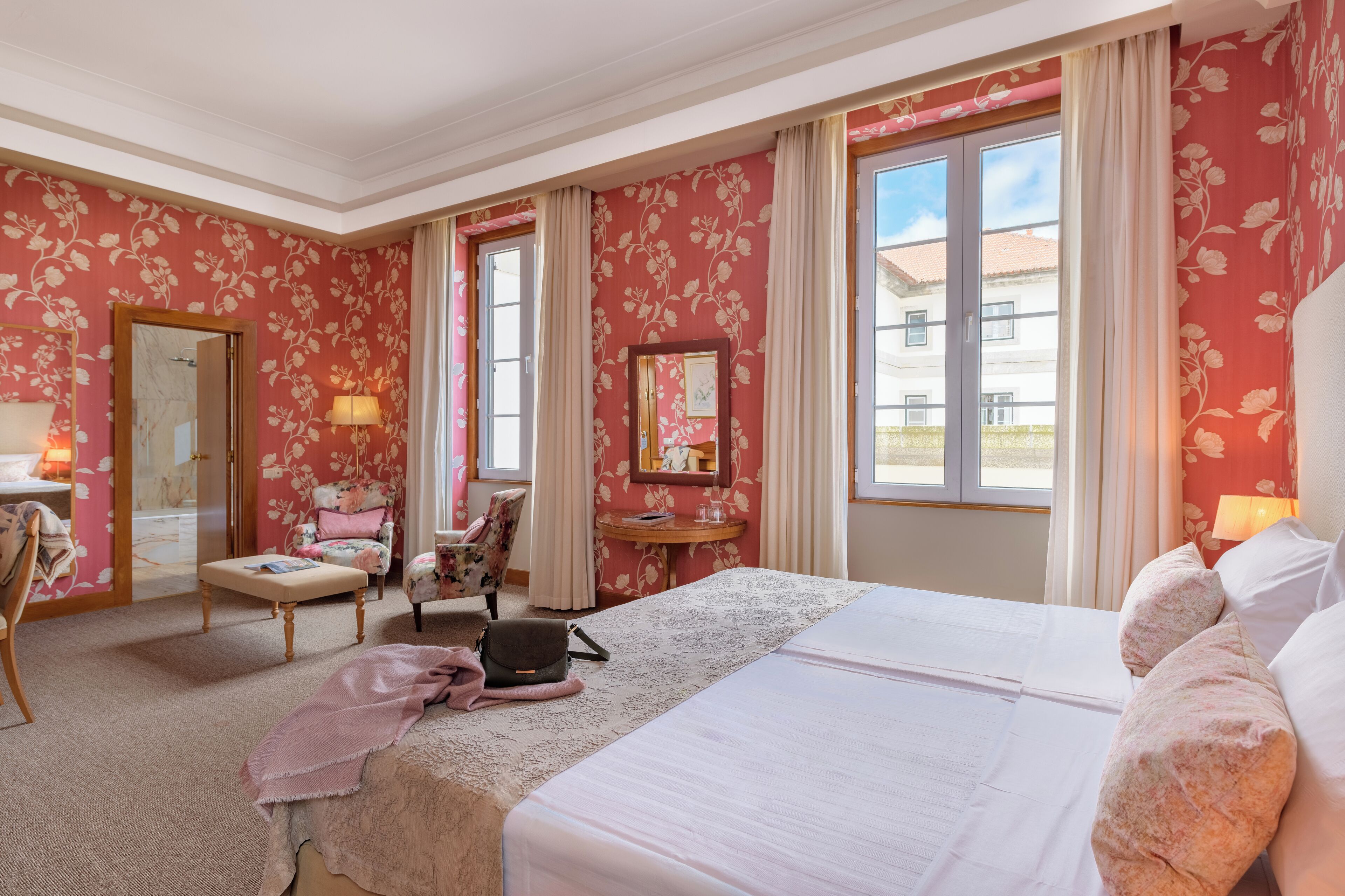 Une chambre d’hôtel avec un papier peint à motifs floraux, un grand lit, un coin salon avec un fauteuil et une table, et une vue sur l’extérieur par la fenêtre.