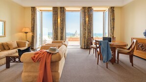 Une chambre d’hôtel équipée d’un canapé, d’un fauteuil, d’une table à manger et de chaises. De grandes fenêtres offrent une vue sur le paysage urbain.