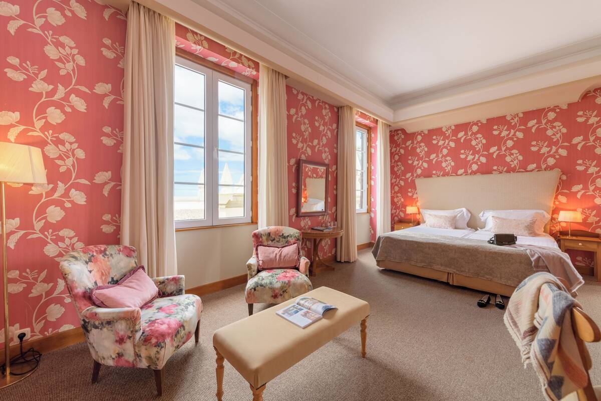 Habitació d'hotel amb paper pintat floral, un llit, dues butaques, una panxa i una tauleta de nit amb una làmpada, i una finestra amb cortines.