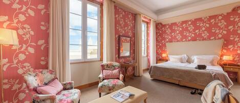 A hotel room with floral wallpaper, a bed, two armchairs, a bench, a bedside table with a lamp, and a window with curtains.
