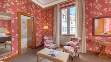 Una habitación con papel tapiz floral, una cama, un banco con una revista, dos sillones florales, una mesa con una lámpara y un ventanal grande.