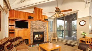 A cozy living room with a stone fireplace, wooden entertainment center, and large windows offering a view of the outdoors. - Forest Condominiums by Vail Resorts (Keystone)
