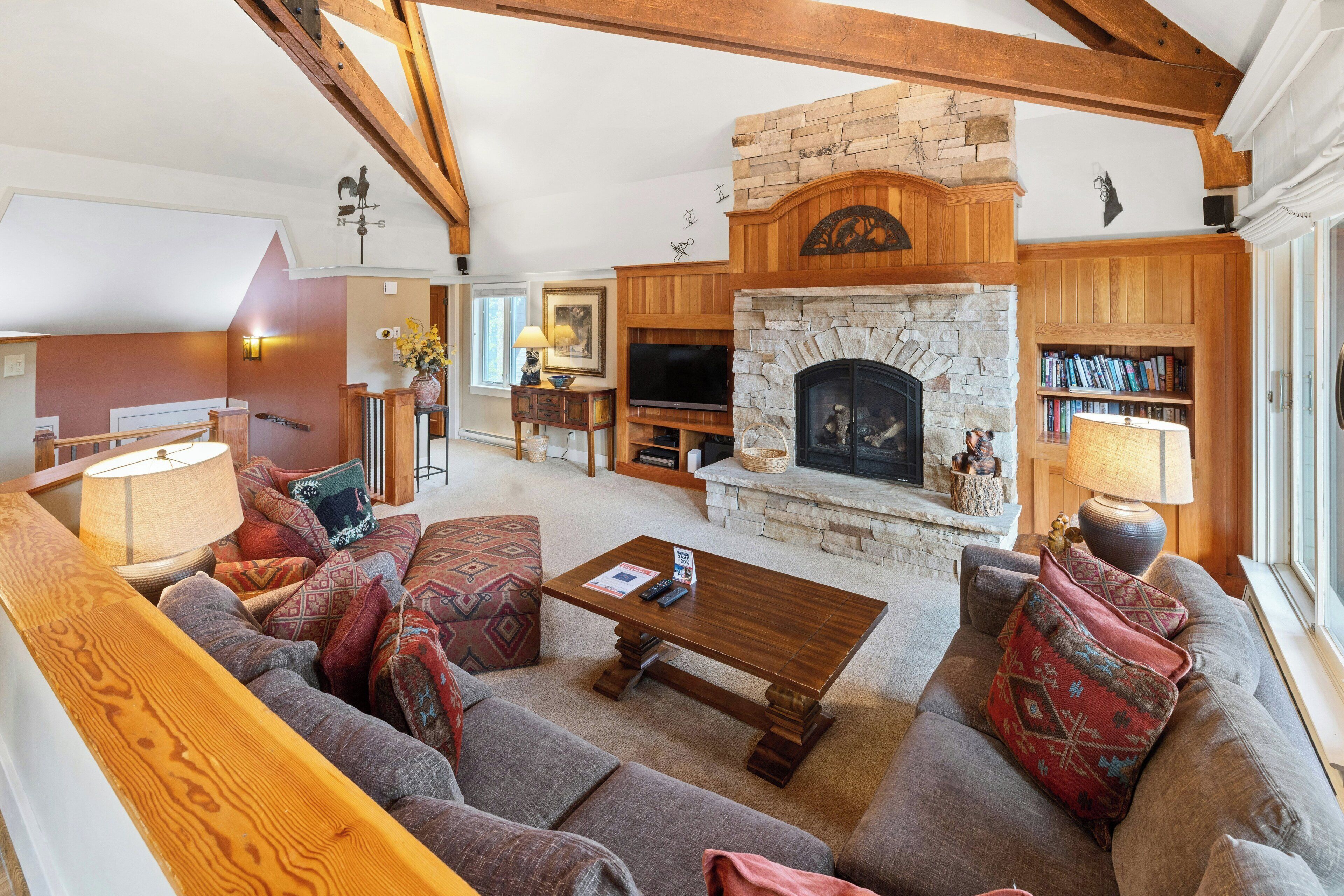A cozy living room with a stone fireplace, wooden beams, a sofa set, and a bookshelf.