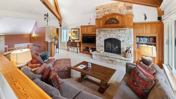 A cozy living room with a stone fireplace, wooden beams, a sofa set, and a bookshelf.