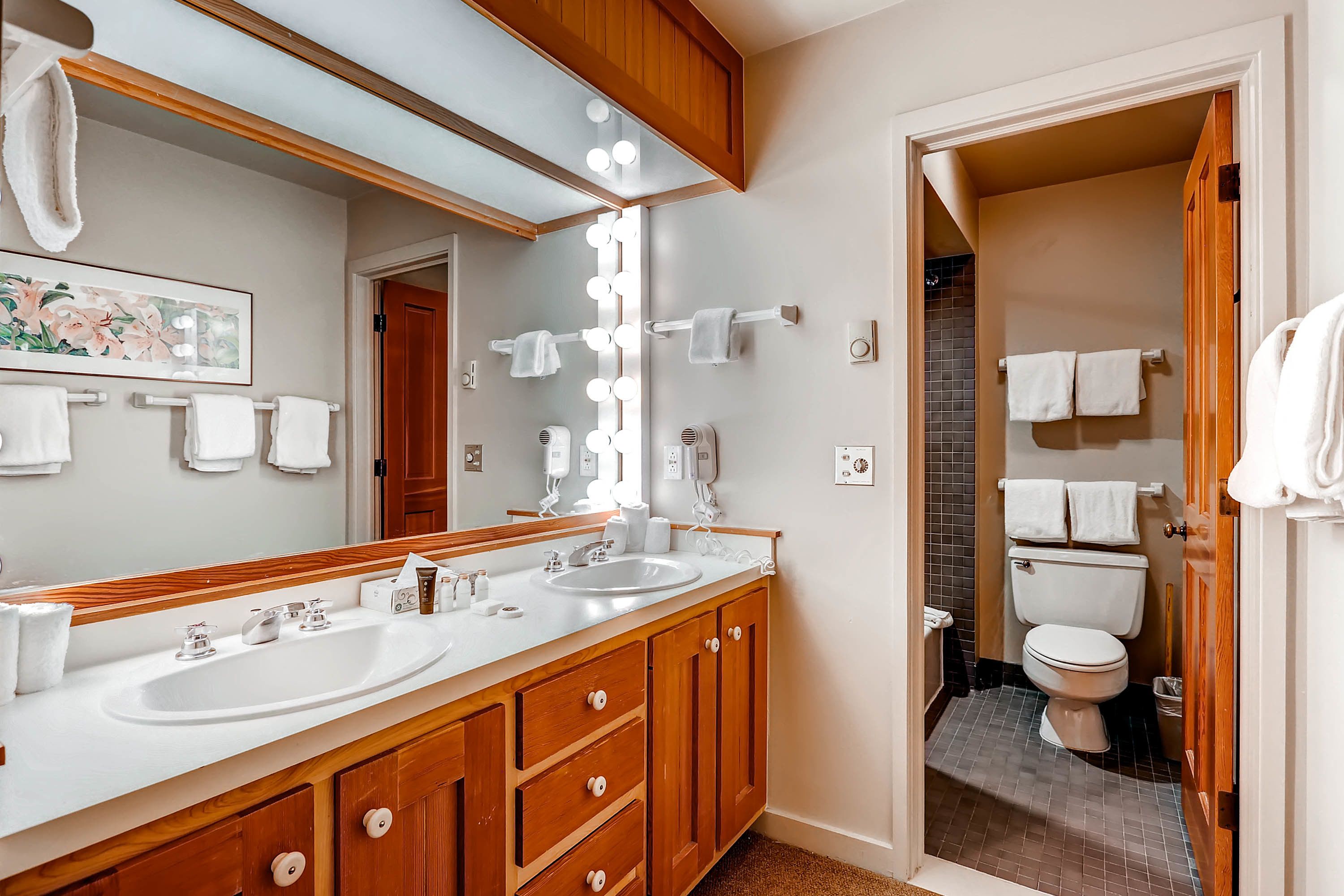 Une salle de bain avec un meuble-lavabo en bois, deux vasques, un grand miroir avec éclairage intégré et des toilettes visibles à travers une porte ouverte.
