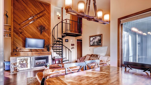 A spacious living room with a wooden floor, a fireplace, a television, a dining area with a table and chairs, and a staircase leading to an upper level. - Forest Condominiums by Vail Resorts (Keystone)