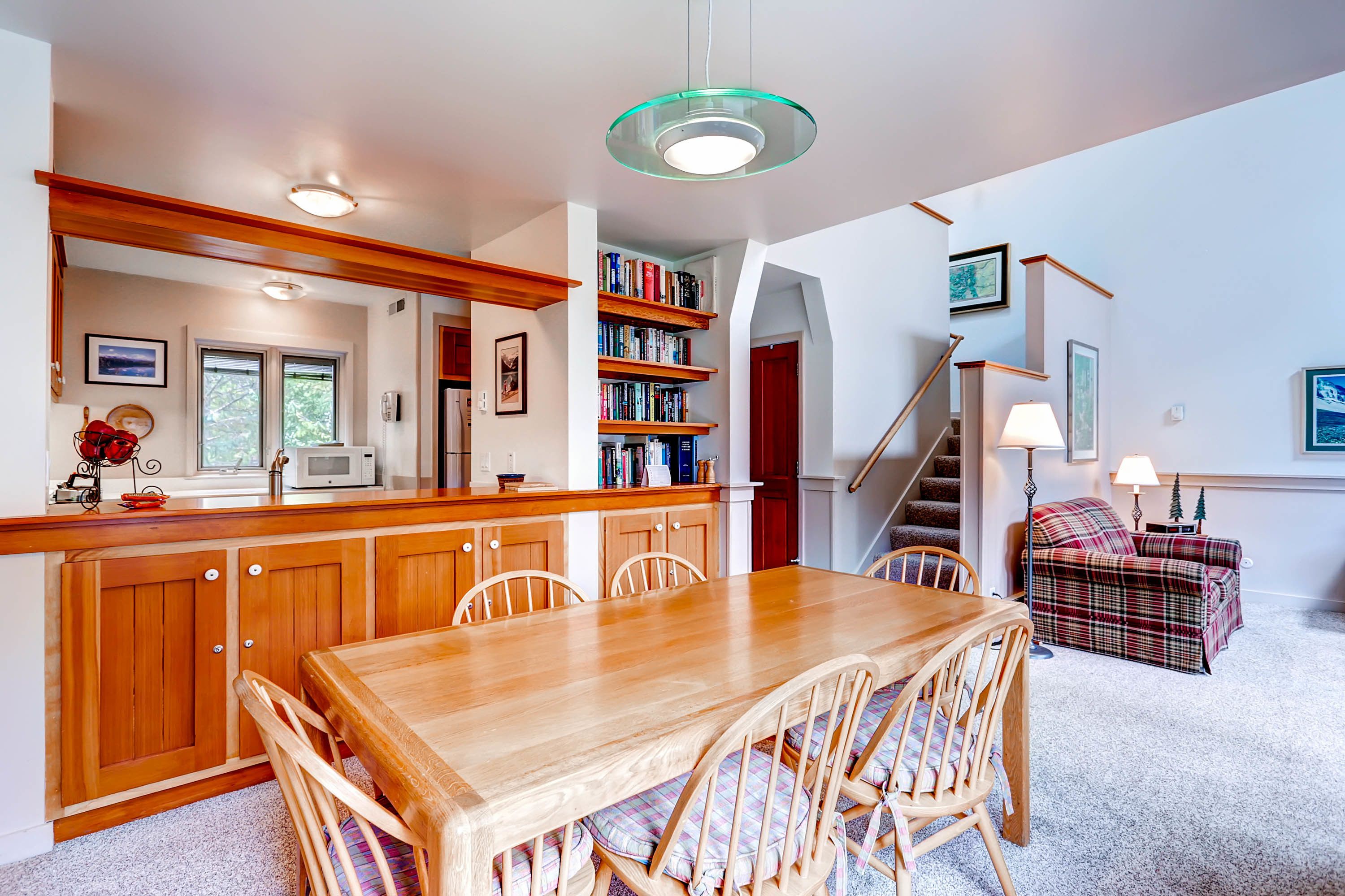 A spacious kitchen with wooden cabinets, a dining table, and chairs. There is a staircase leading to another level, and a bookshelf filled with books.