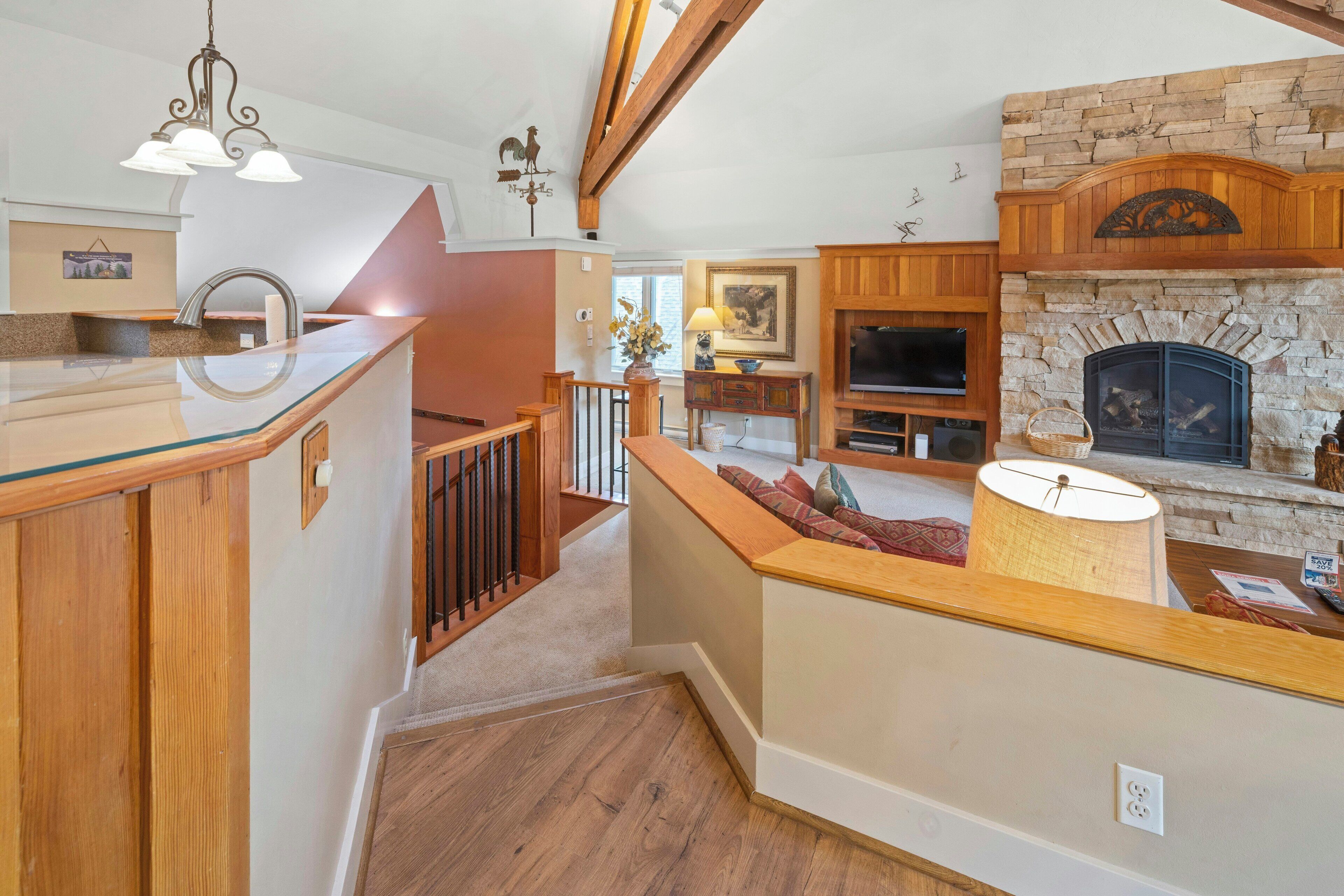 A spacious living area with a stone fireplace, wooden beams, a kitchen with a glass island, and a staircase leading to another level.