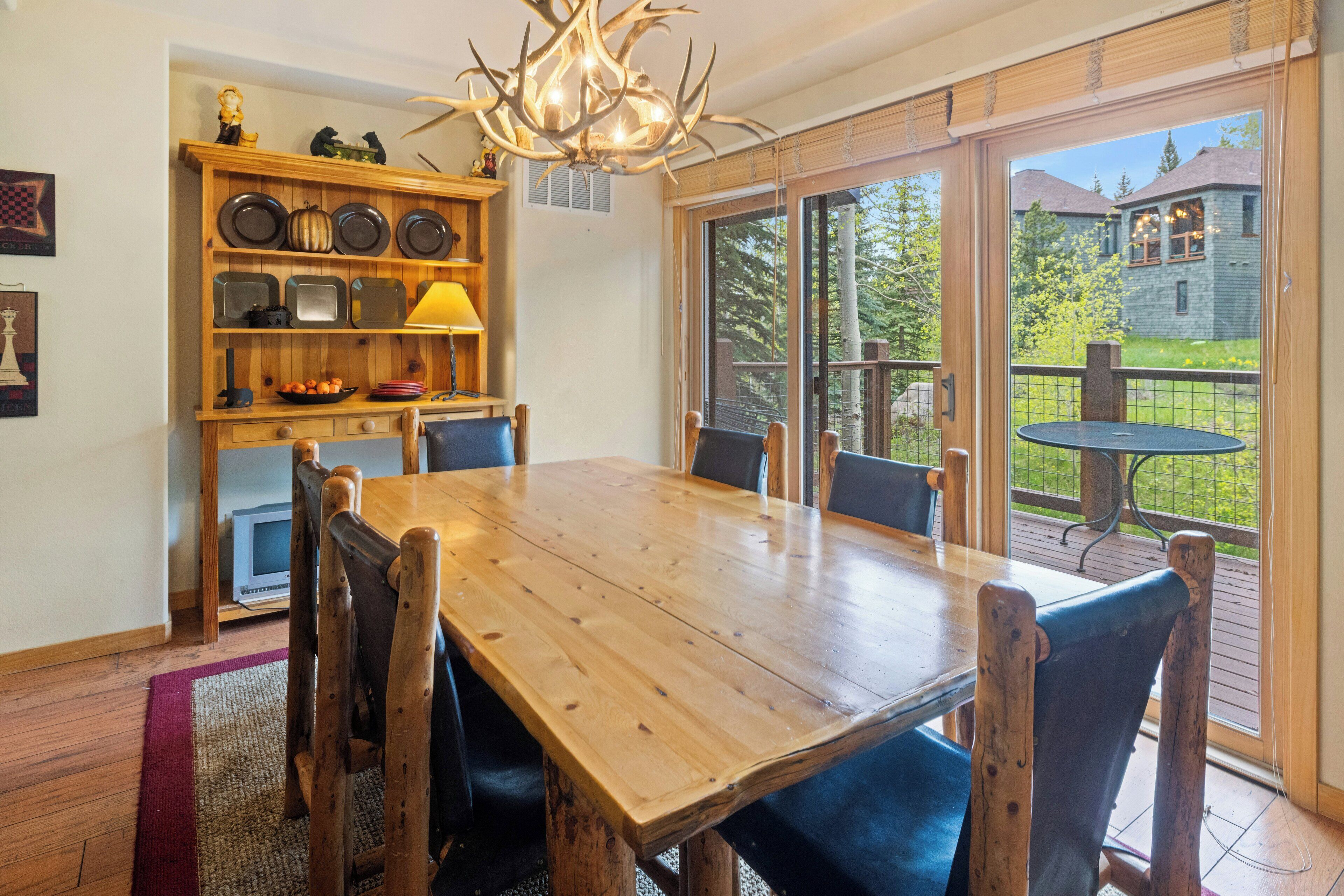 A wooden dining table with chairs, a chandelier, and a kitchen area with shelves and appliances.