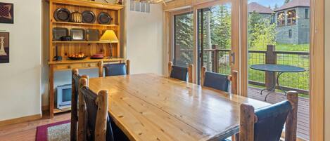A wooden dining table with chairs, a chandelier, and a kitchen area with shelves and appliances.