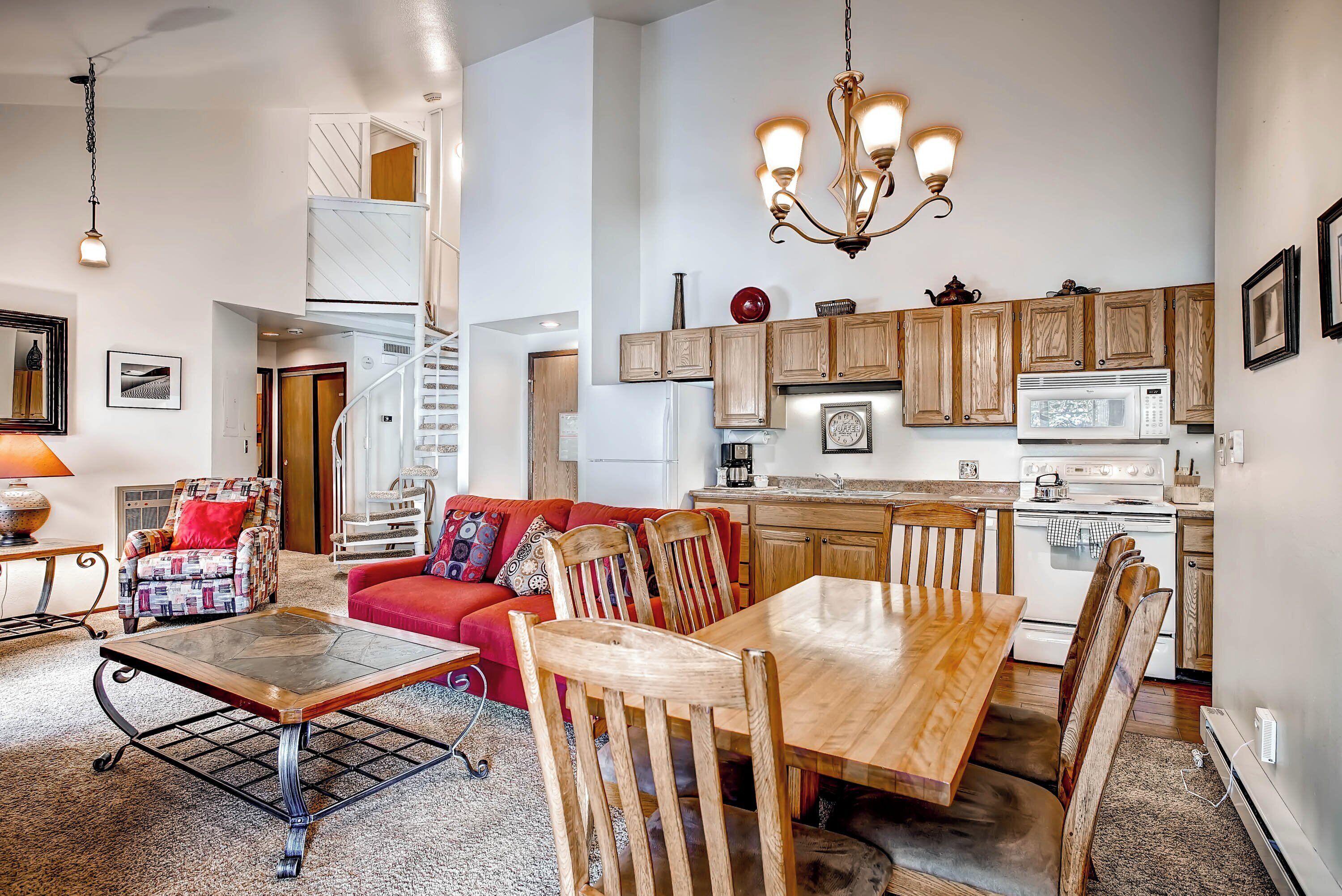 A spacious living area with a red sofa, wooden dining table, and chairs, a kitchen with wooden cabinets and a white stove, and a staircase leading to an upper level.