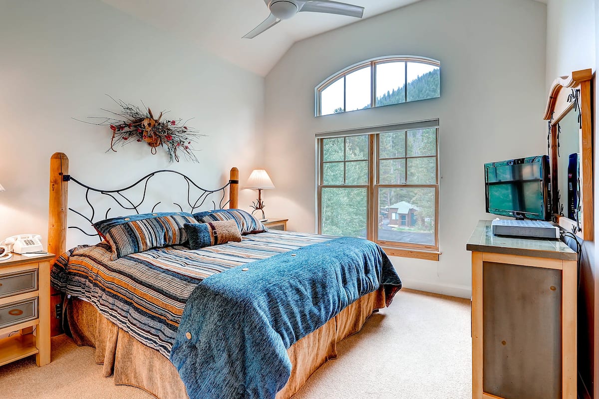 A bedroom with a bed, nightstands, a desk, a computer, and a large window with a view of trees.