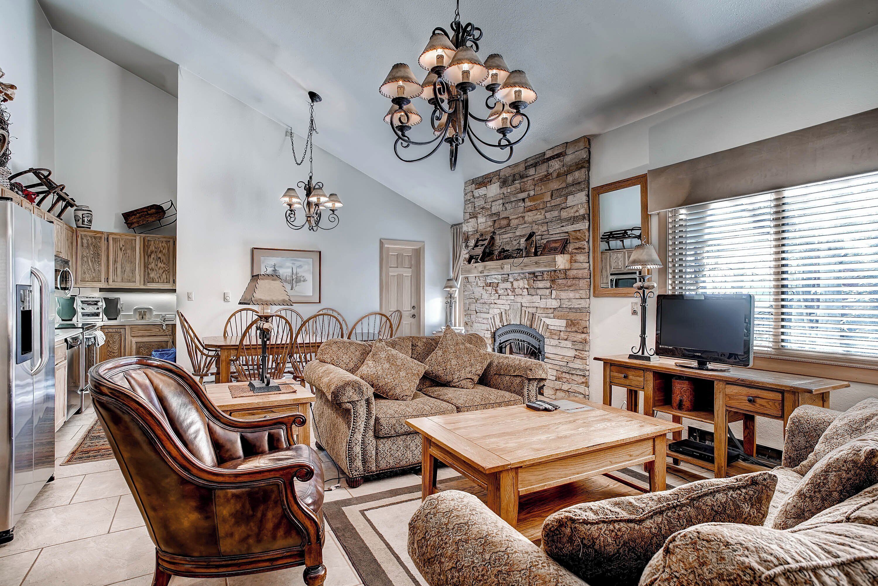 A spacious living room with a sofa, armchairs, a coffee table, and a TV stand. There is a fireplace, a chandelier, and a stone wall.