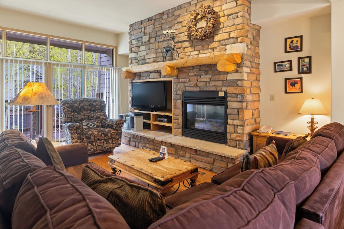 A living room with a stone fireplace, comfortable seating, a television, and a large window.