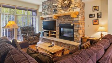 A living room with a stone fireplace, comfortable seating, a television, and a large window.