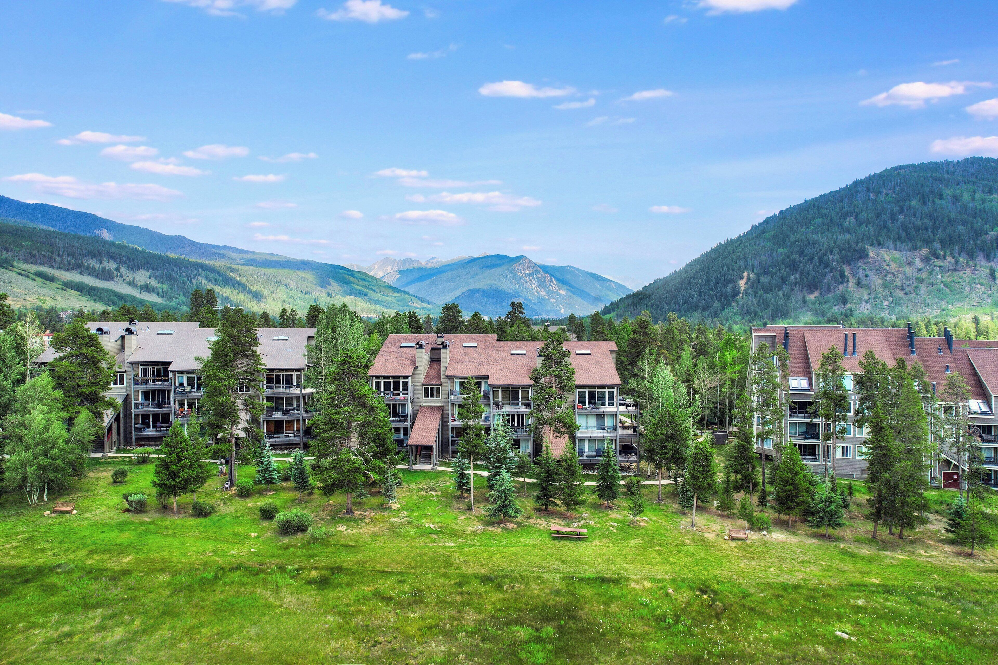 A multi-story building with a flat roof, surrounded by a grassy area and trees, set against a mountainous backdrop.