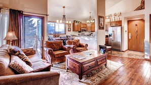 A spacious living room with a large window, leather sofas, a wooden coffee table, and a kitchen in the background. - Forest Condominiums by Vail Resorts (Keystone)