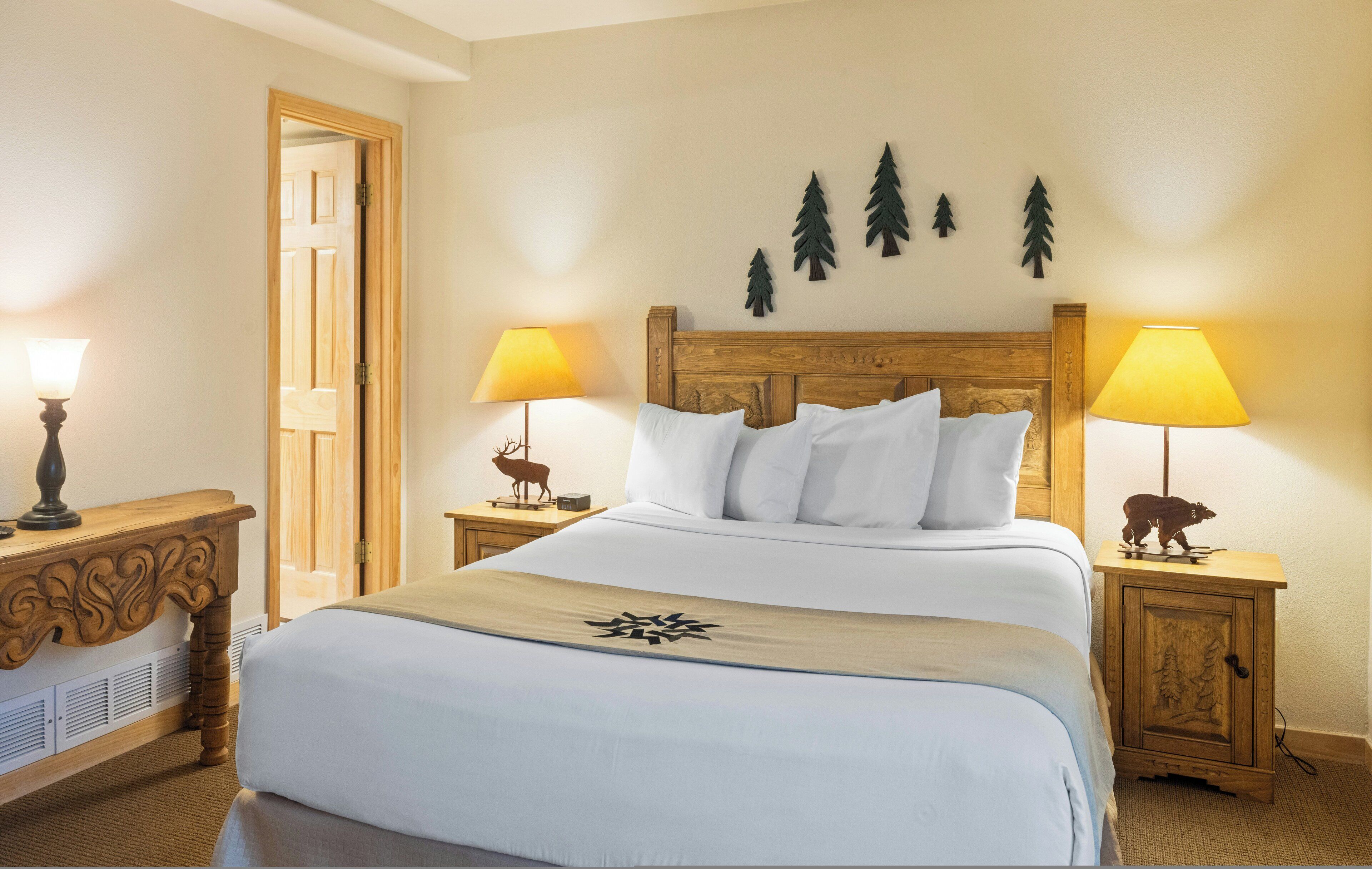 Une chambre d’hôtel bien aménagée, avec un grand lit, deux lampes de chevet, une table de chevet en bois avec une lampe, et une porte donnant sur une autre pièce.