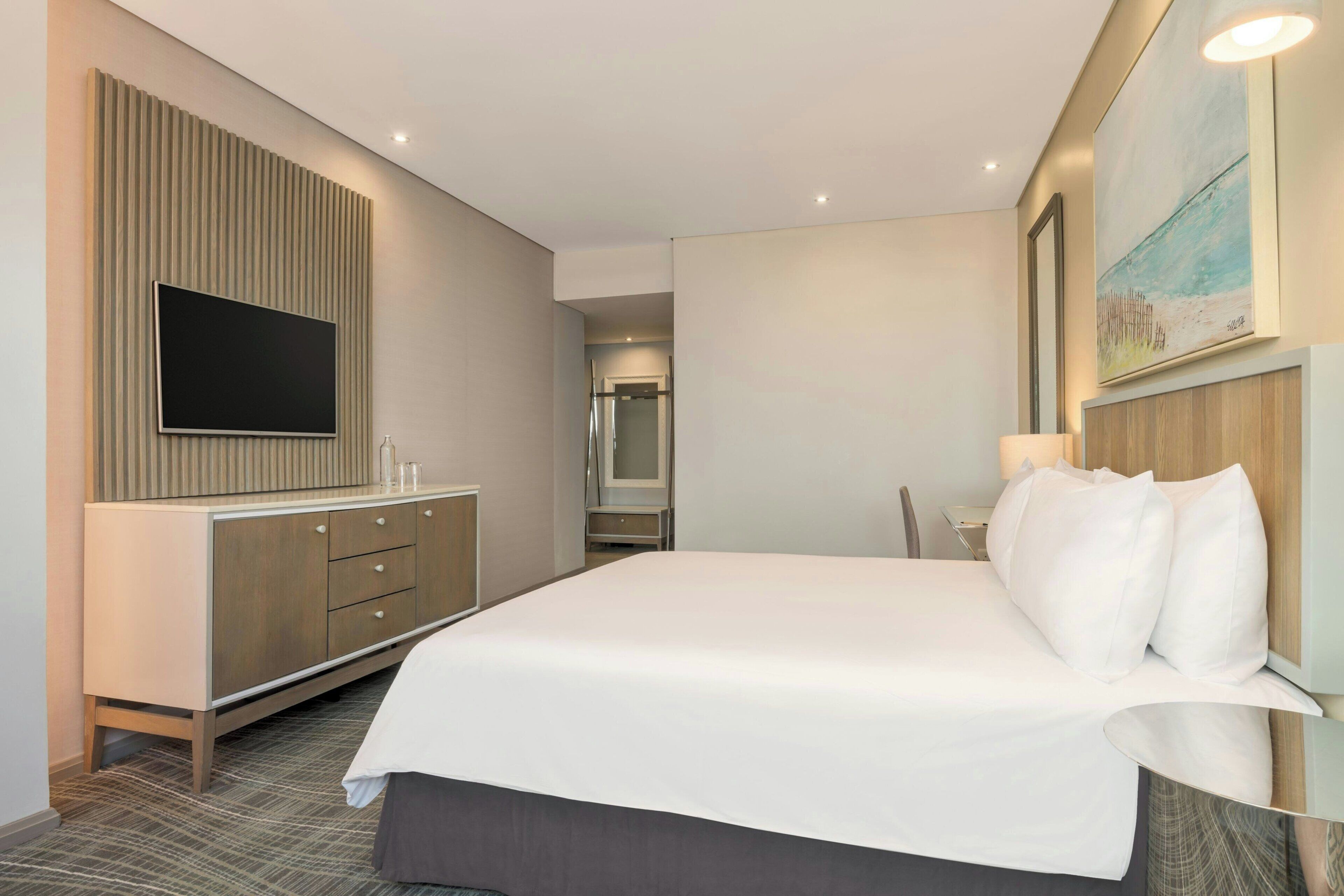A hotel room with a large bed, a flat-screen TV mounted on a wooden panel, a desk with a chair, and a painting on the wall.