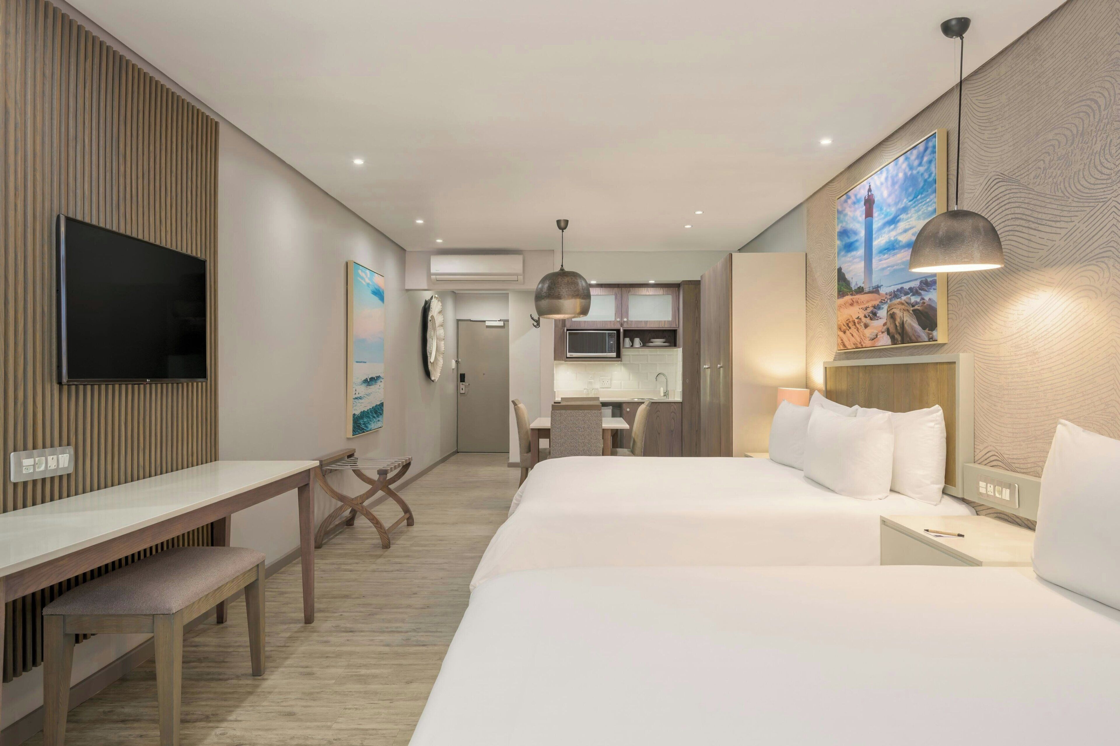 A hotel room with a bed, bedside table, desk, and a TV. There is a kitchenette in the background.