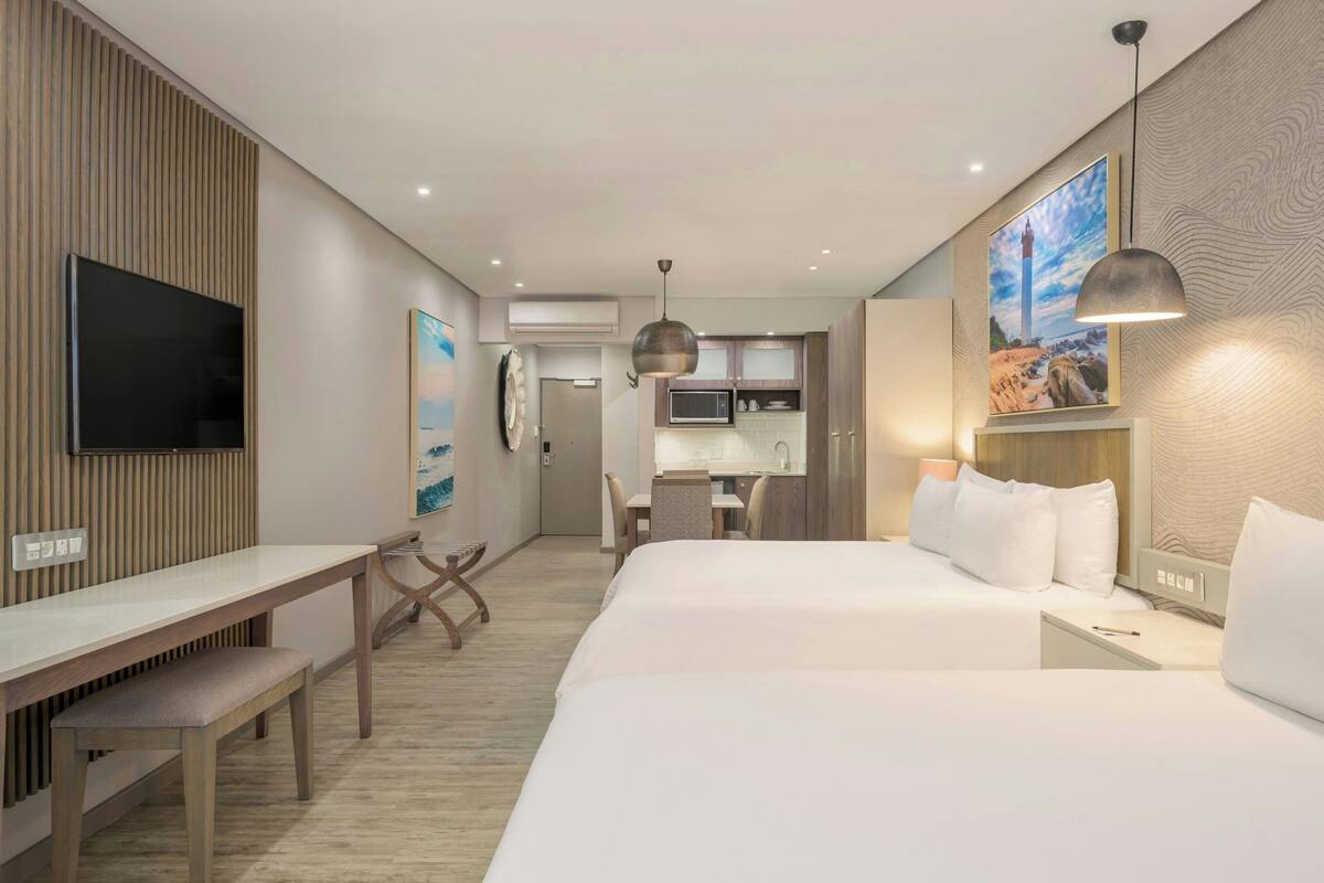 Une chambre d’hôtel avec un lit, une table de chevet, un bureau et un téléviseur. On aperçoit un coin cuisine au fond de la pièce.