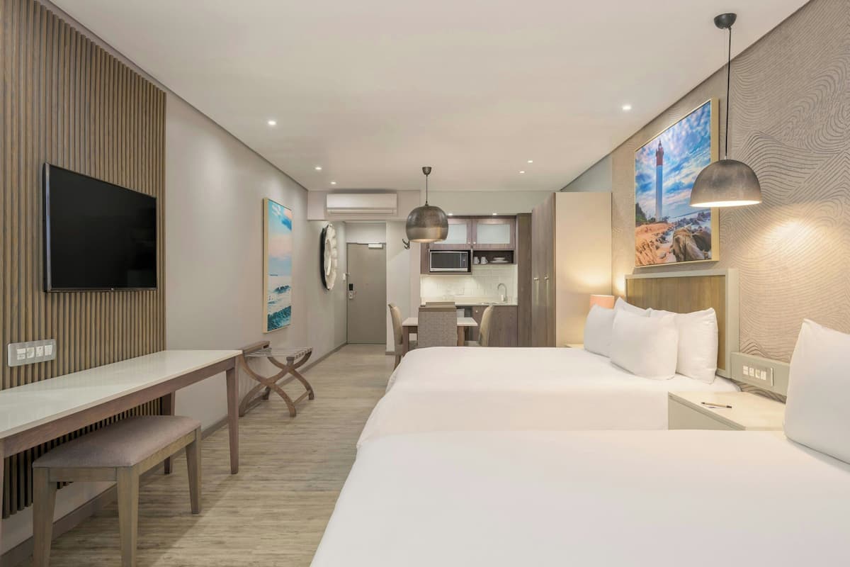 A hotel room with a bed, bedside table, desk, and a TV. There is a kitchenette in the background.