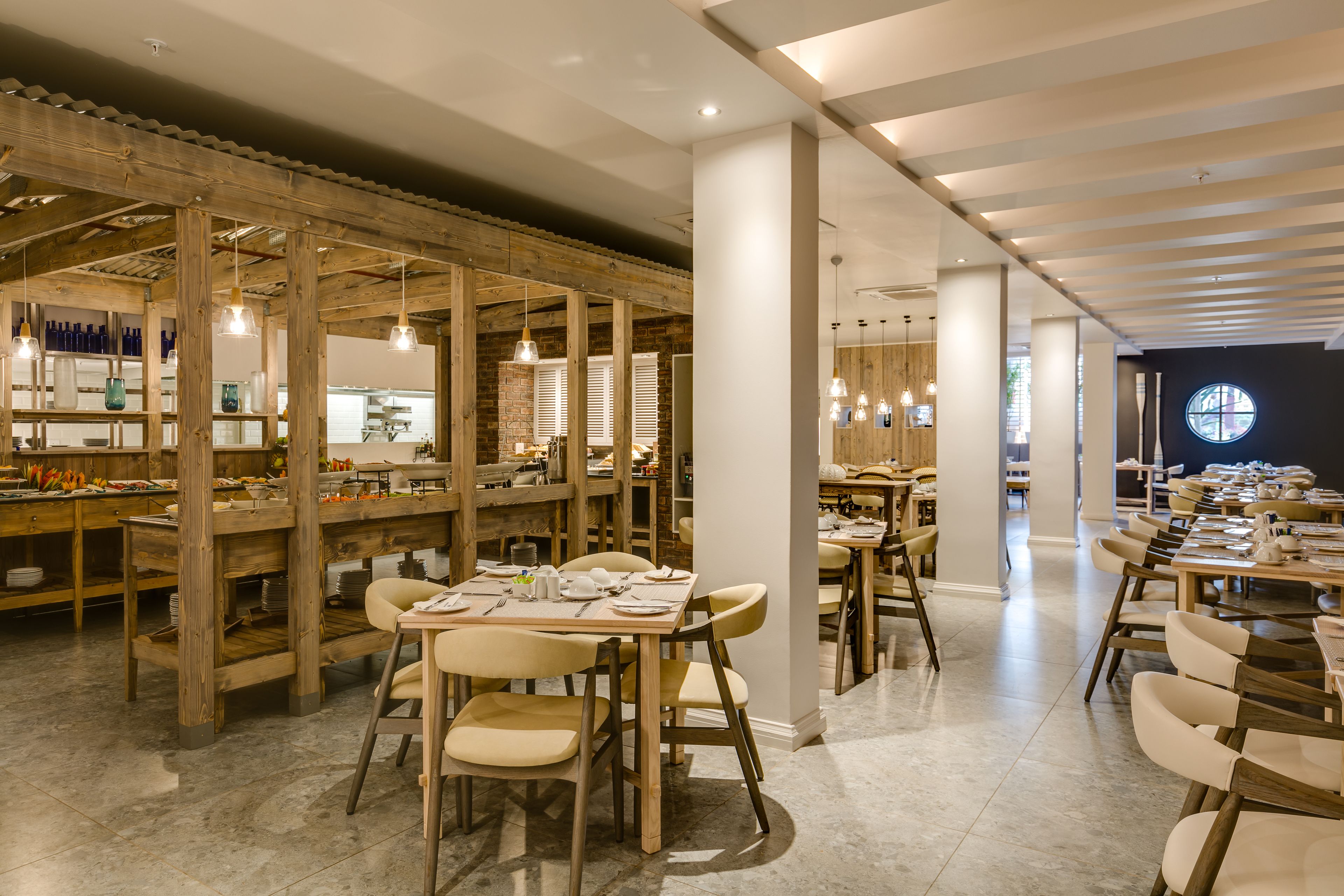 Un intérieur de restaurant moderne, avec des poutres en bois, des piliers blancs et un mélange de tables rondes et rectangulaires.