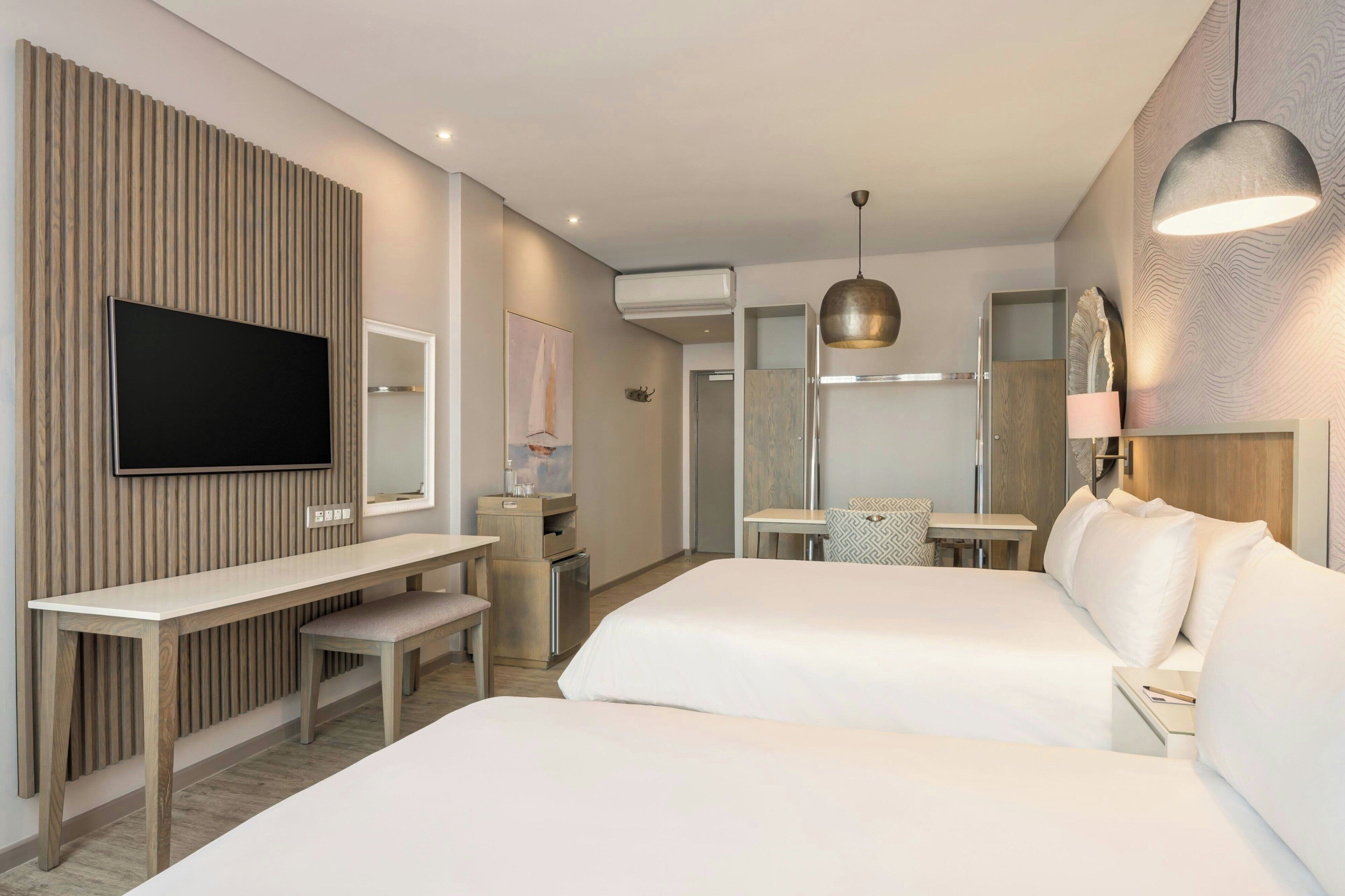 A modern hotel room with a flat-screen TV, two beds, a desk, and a large mirror.