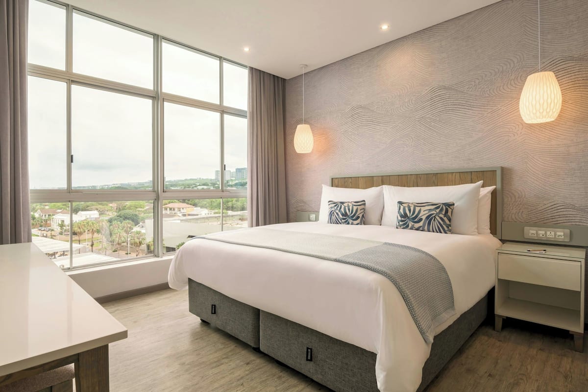 Une chambre d’hôtel moderne avec une grande fenêtre, un lit doté d’une tête de lit en bois, une table de chevet et une lampe fixée au mur.